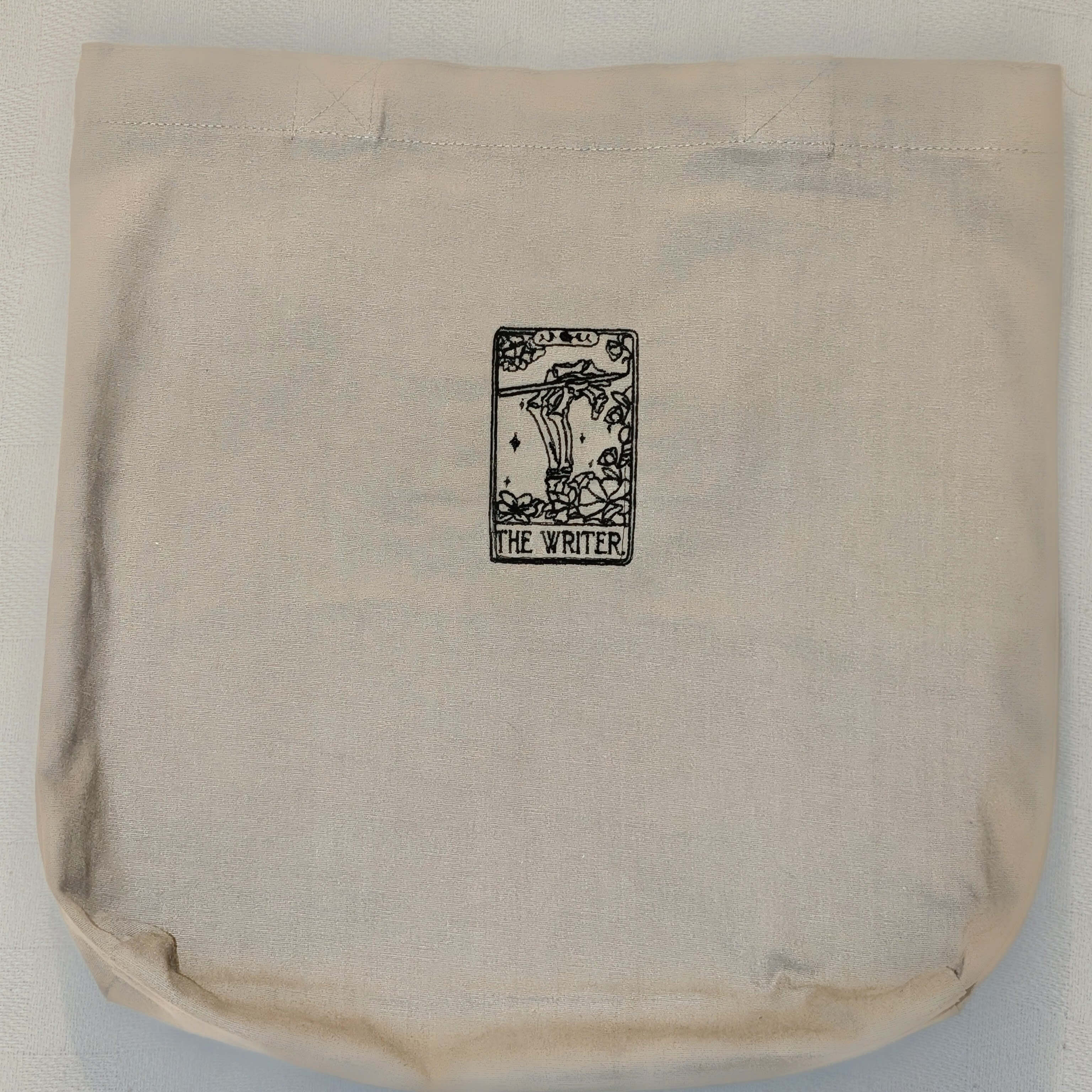Cream-colored tote bag showcasing a tarot-inspired embroidery of a skeleton sitting at a desk, labeled The Writer.